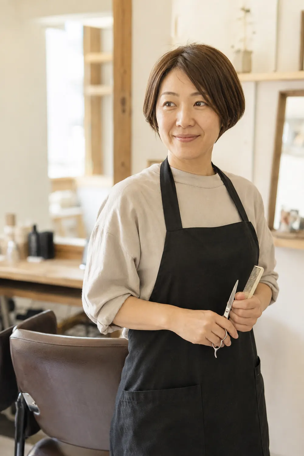黒いエプロンをつけ、はさみとコームを持って立つショートボブの女性スタイリスト